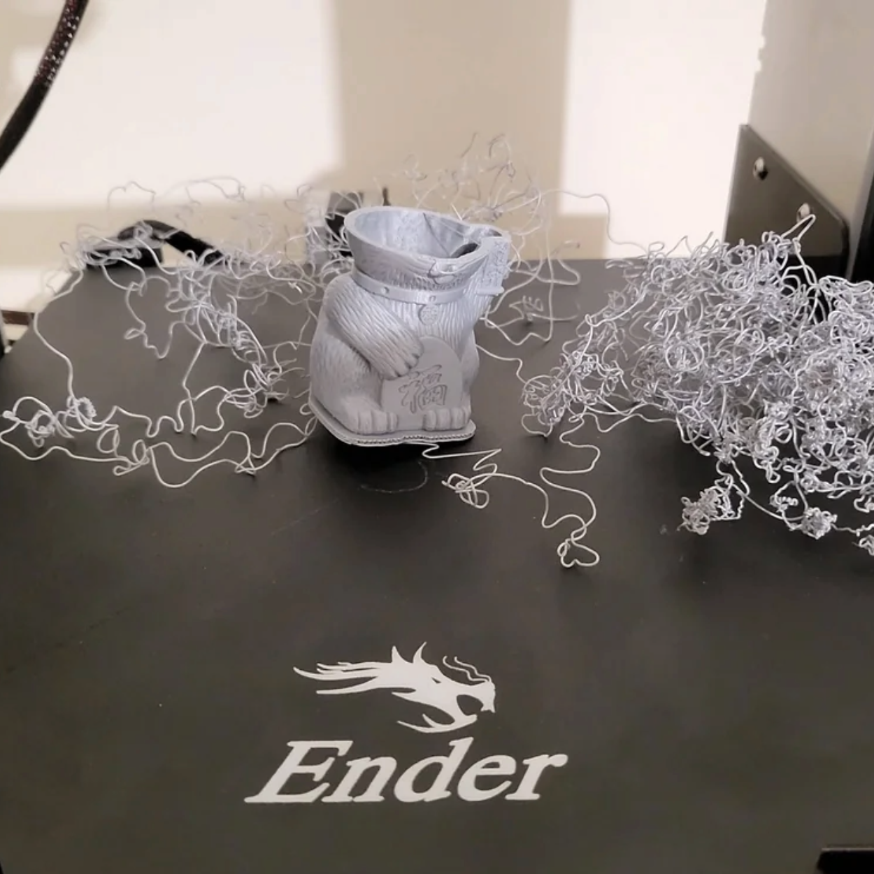 Mislukte print van een grijs figuurtje op een Ender-printbed, omringd door slierten filament
