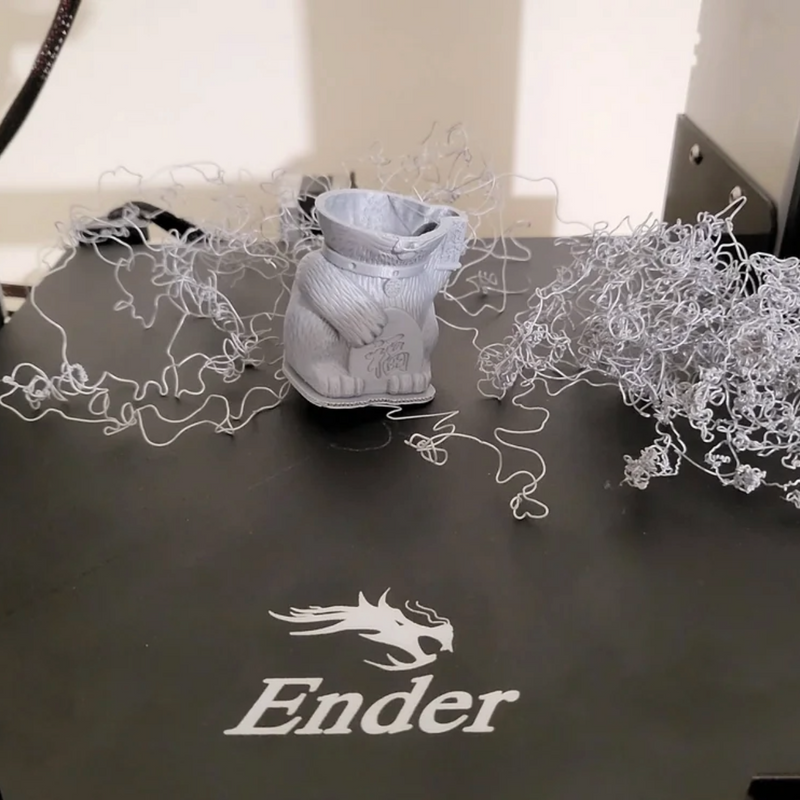 Mislukte print van een grijs figuurtje op een Ender-printbed, omringd door slierten filament